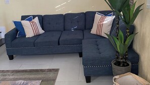 A living room with a dark blue sectional sofa, a large potted plant, a woven basket, and two framed artworks on the wall.