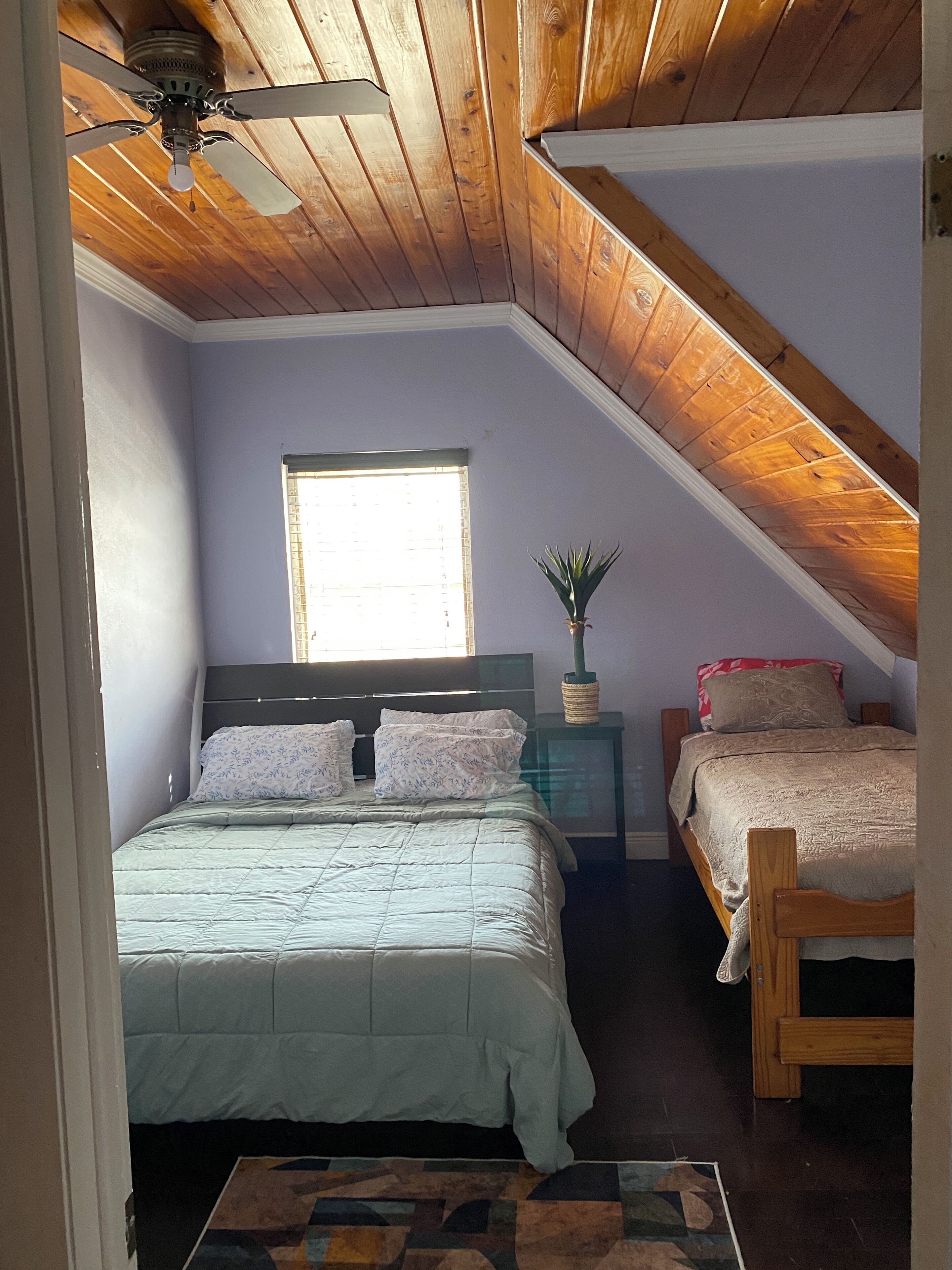 A bedroom with a sloped ceiling, a large bed, a smaller bed, a window, and a ceiling fan.