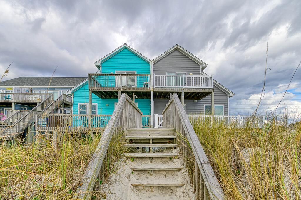 A beachfront property with a wooden staircase leading to a balcony, surrounded by tall grass and under a cloudy sky.