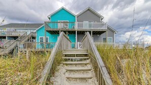 Een strandpand met een houten trap naar een balkon, omgeven door hoog gras en onder een bewolkte lucht.