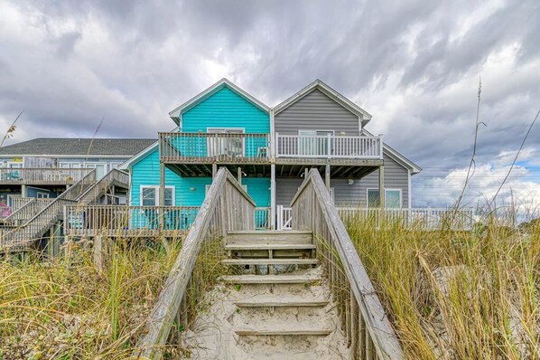 A beachfront property with a wooden staircase leading to a balcony, surrounded by tall grass and under a cloudy sky.