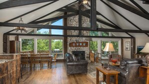A spacious living room with a stone fireplace, wooden beams, and a large window overlooking greenery. - Historic Beech Mountain Lodge | Hot Tub | Game Room | Sleeps 19 (Beech Mountain)
