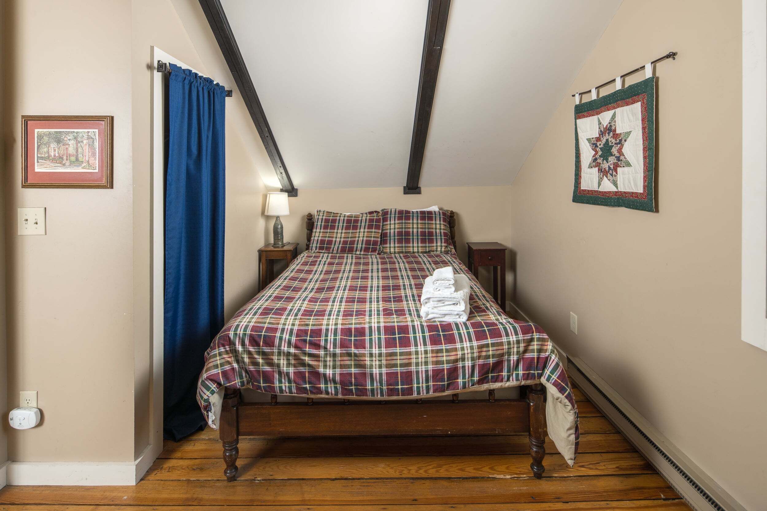 A bedroom with a wooden bed, a blue curtain, a bedside lamp, a framed picture, and a quilt on the wall.