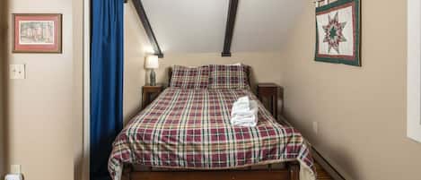 A bedroom with a wooden bed, a blue curtain, a bedside lamp, a framed picture, and a quilt on the wall.