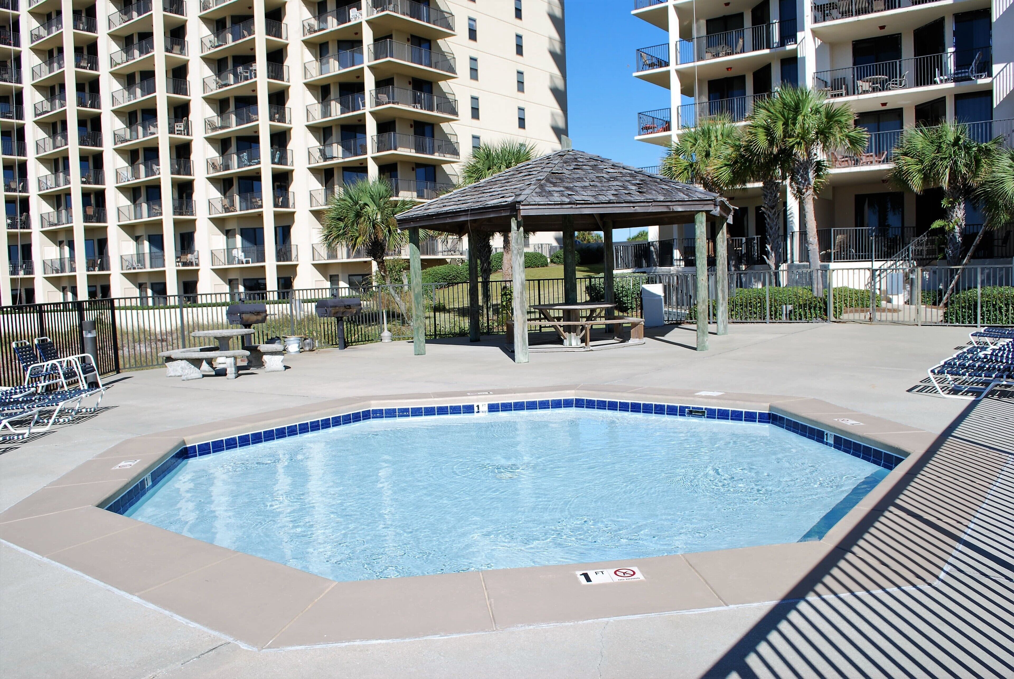 Piscina com espreguiçadeiras e um gazebo, cercada por edifícios altos.