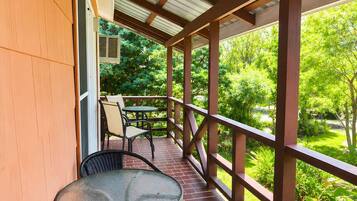 A covered outdoor patio with a table and chairs, a view of greenery, and a wooden railing.