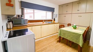 A compact kitchen with a microwave, toaster, and kettle, a dining table with a checkered tablecloth, and a window with a dark blind. - Snowy Mtn Lake Cottage | Pets & Groups | Sleeps 8 (Talbingo)