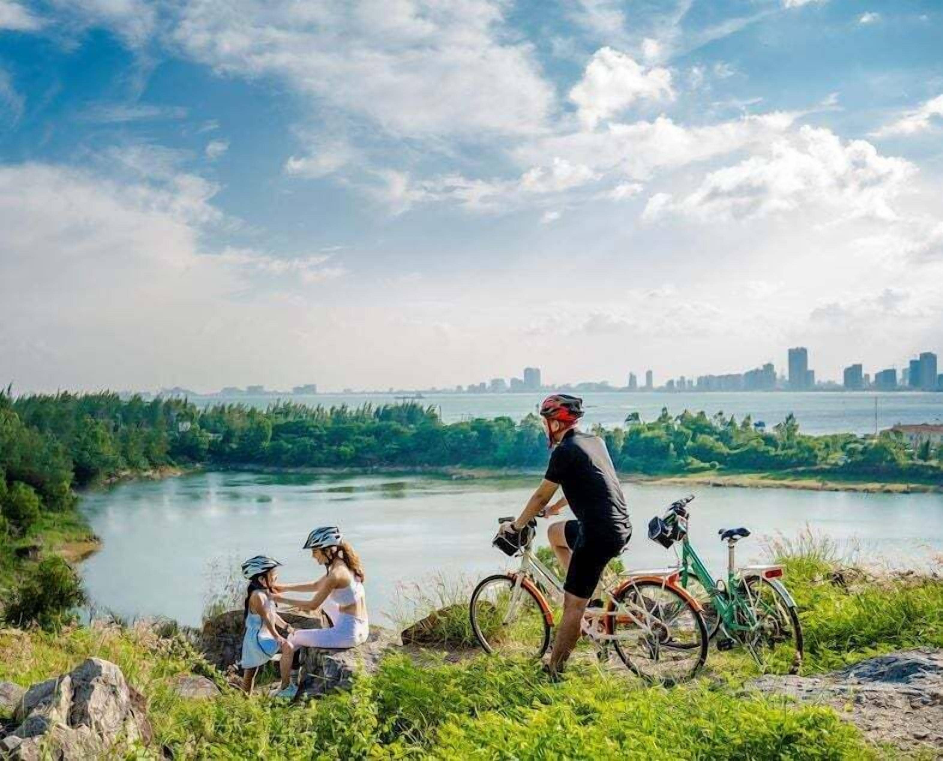 Uitzicht op een rivier met een stadsachtergrond op de achtergrond, een persoon op een fiets en twee kinderen met helmen.