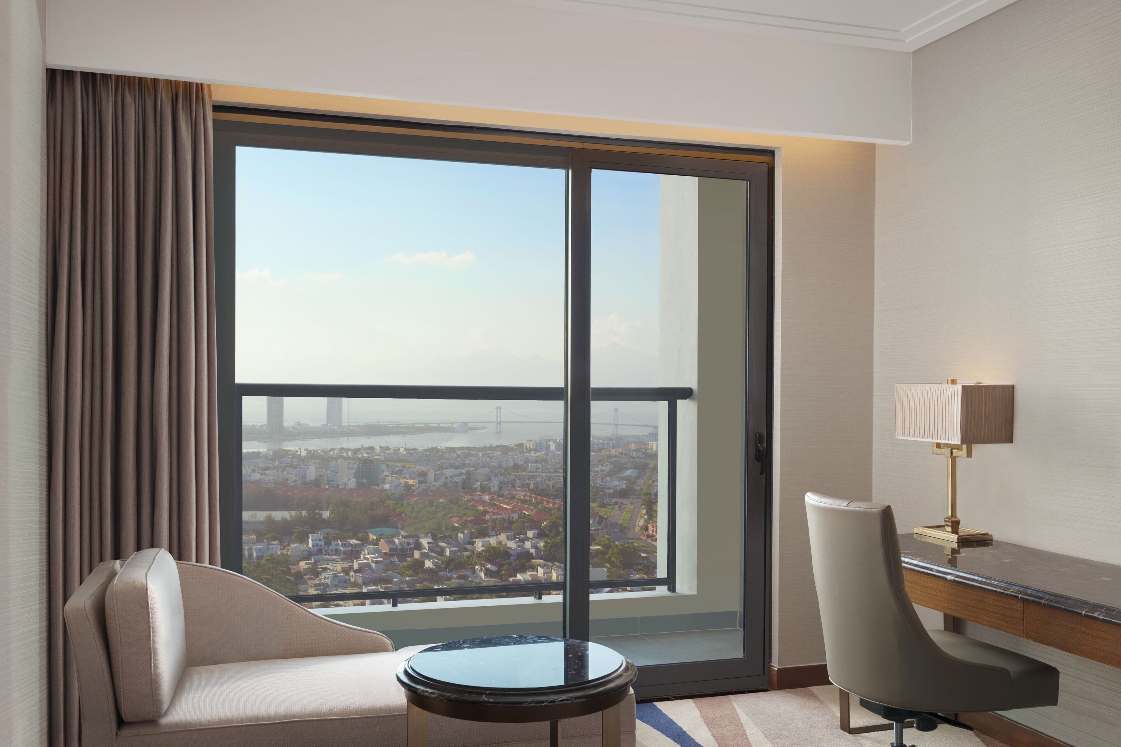 A hotel room with a large window, a sofa, a desk, and a lamp.
