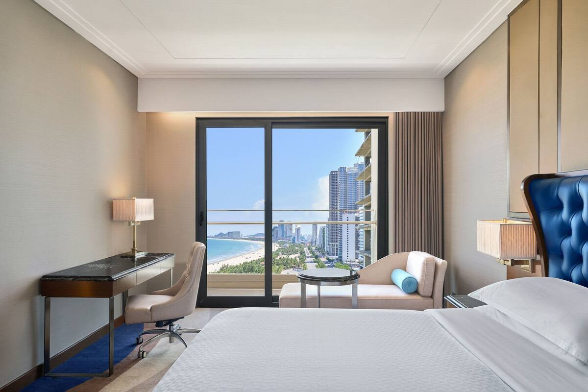 A hotel room with a large bed, a desk, a chair, a small table, and a view of the beach and city skyline.