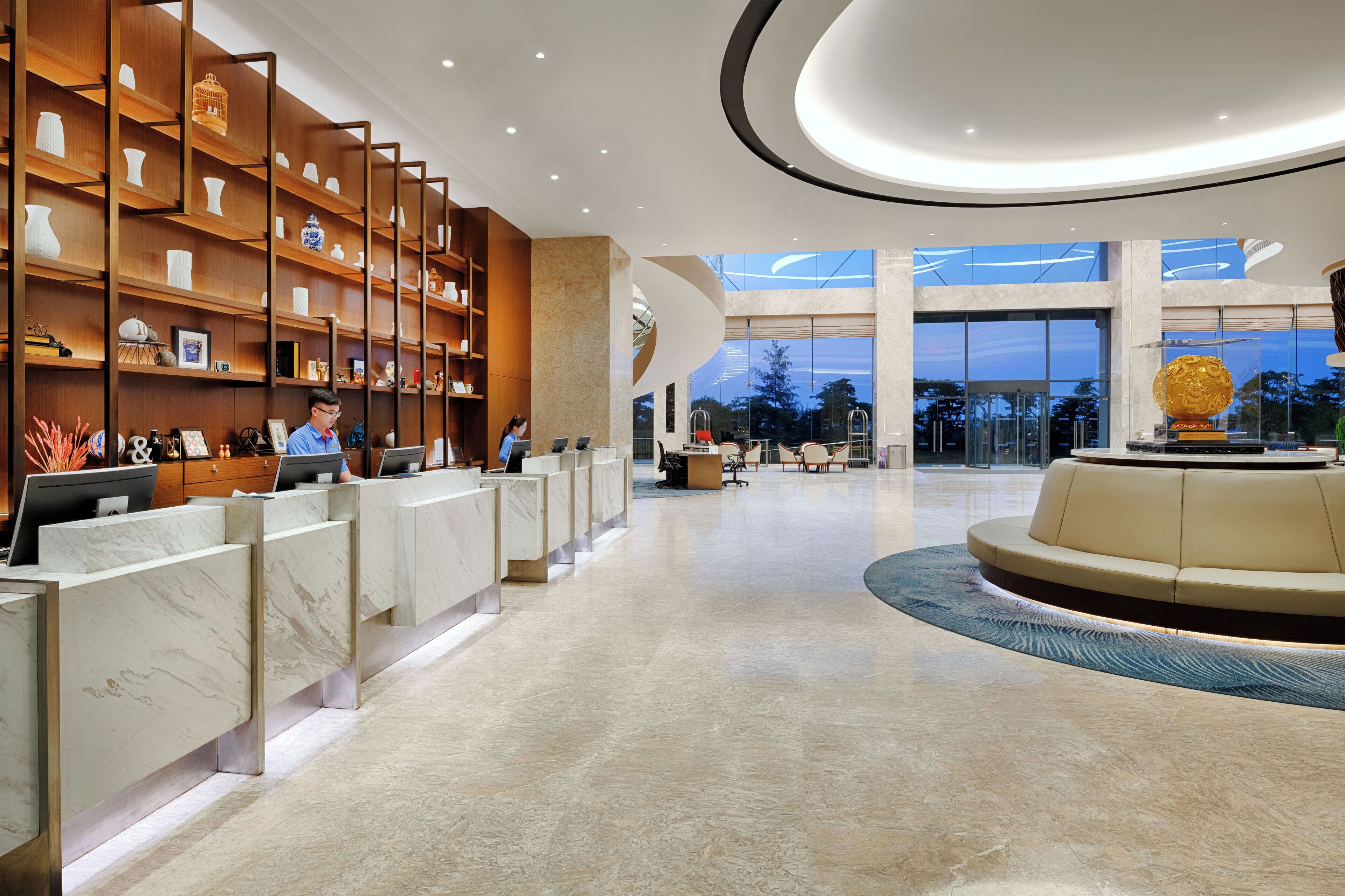 A modern hotel lobby with marble reception counters, a central seating area, and a large window offering a view of the outdoors.