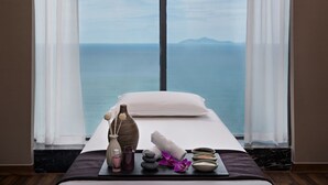 Un cuarto de spa con una cama, una bandeja con artículos de spa, vista al mar y una ventana con cortinas translúcidas.