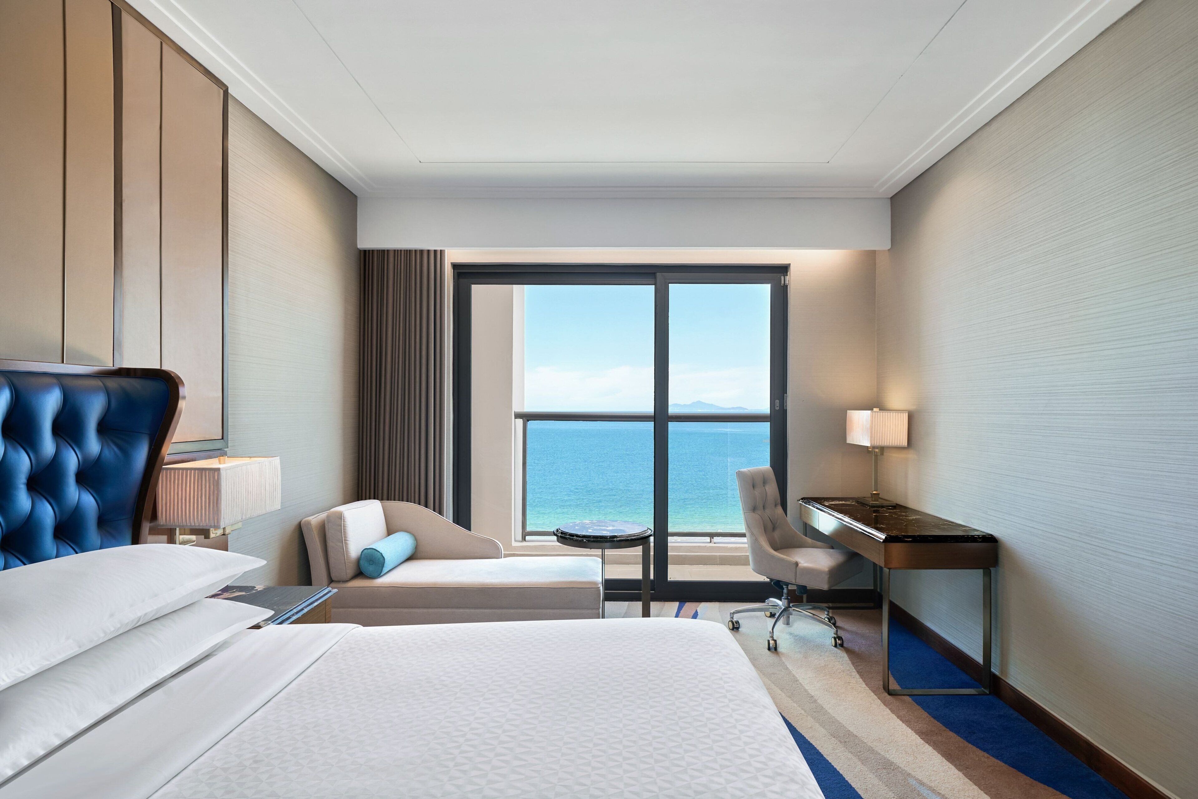 A hotel room with a large bed, a desk, a chair, and a view of the ocean through a large window.
