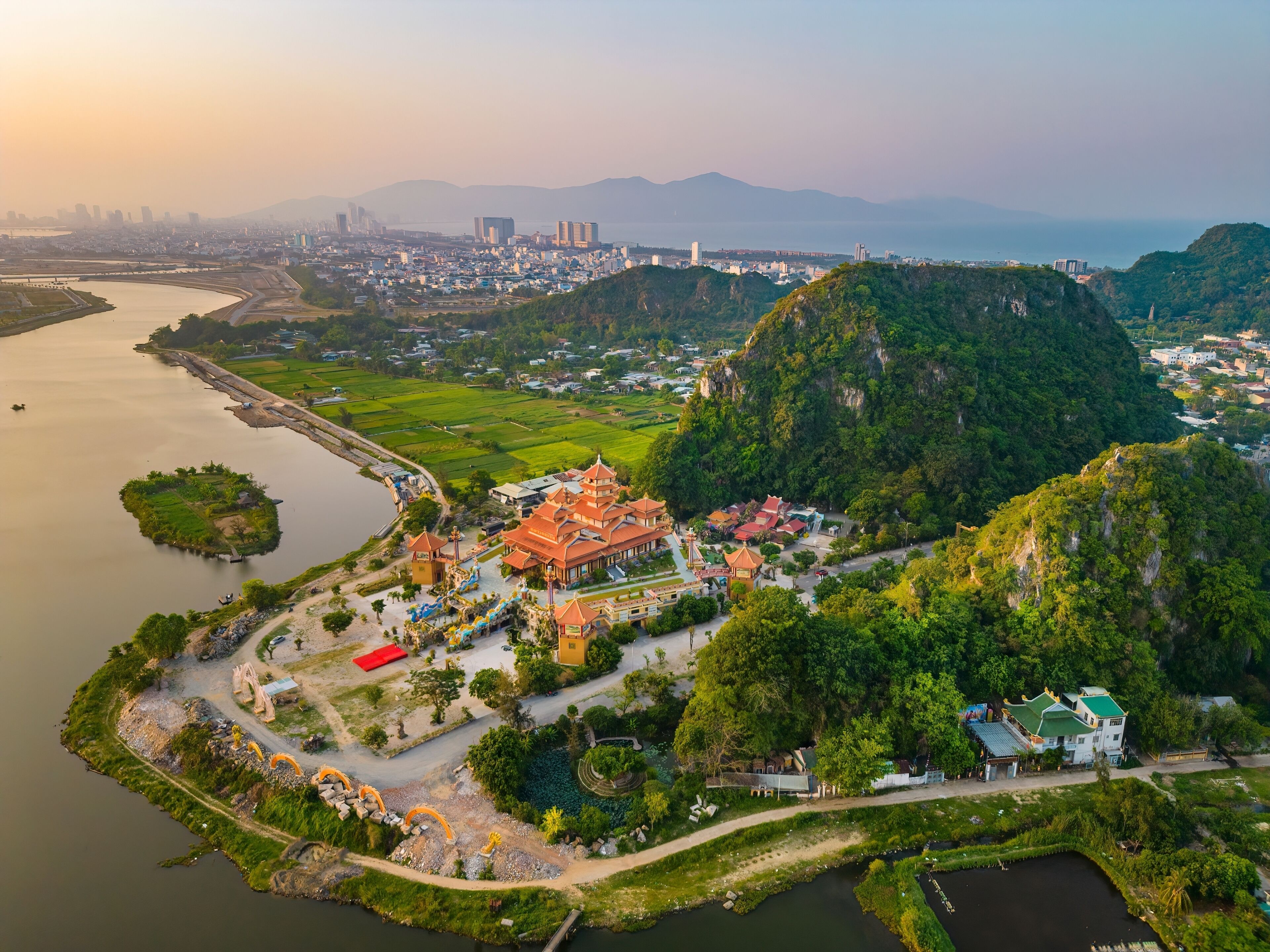 An aerial view of a traditional temple complex with a river, lush greenery, and a distant city skyline.