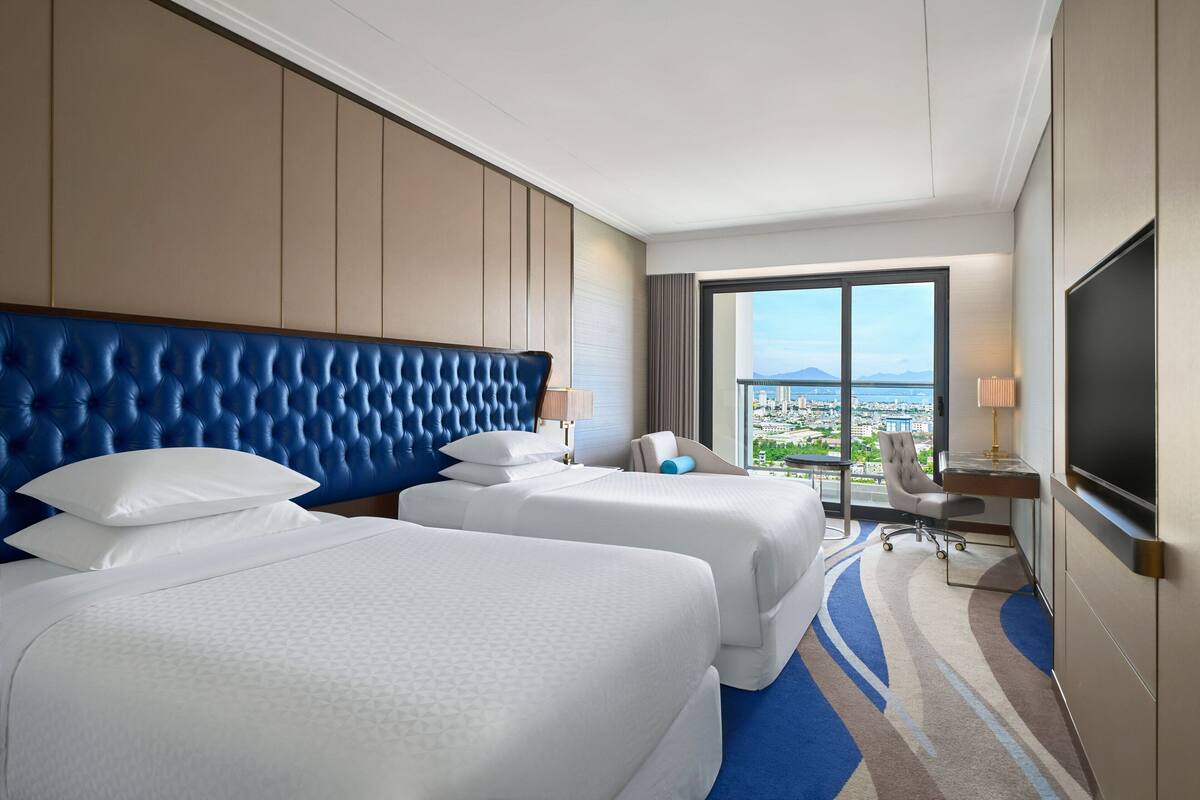 A hotel room with two beds, a TV, a desk, and a view of the city.
