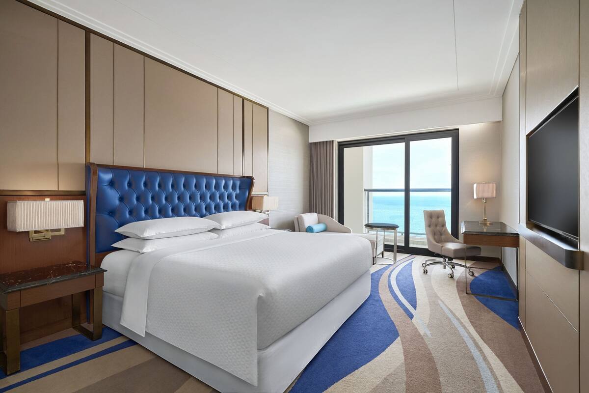 A hotel room with a large bed, a desk, a chair, a TV, and a view of the ocean.