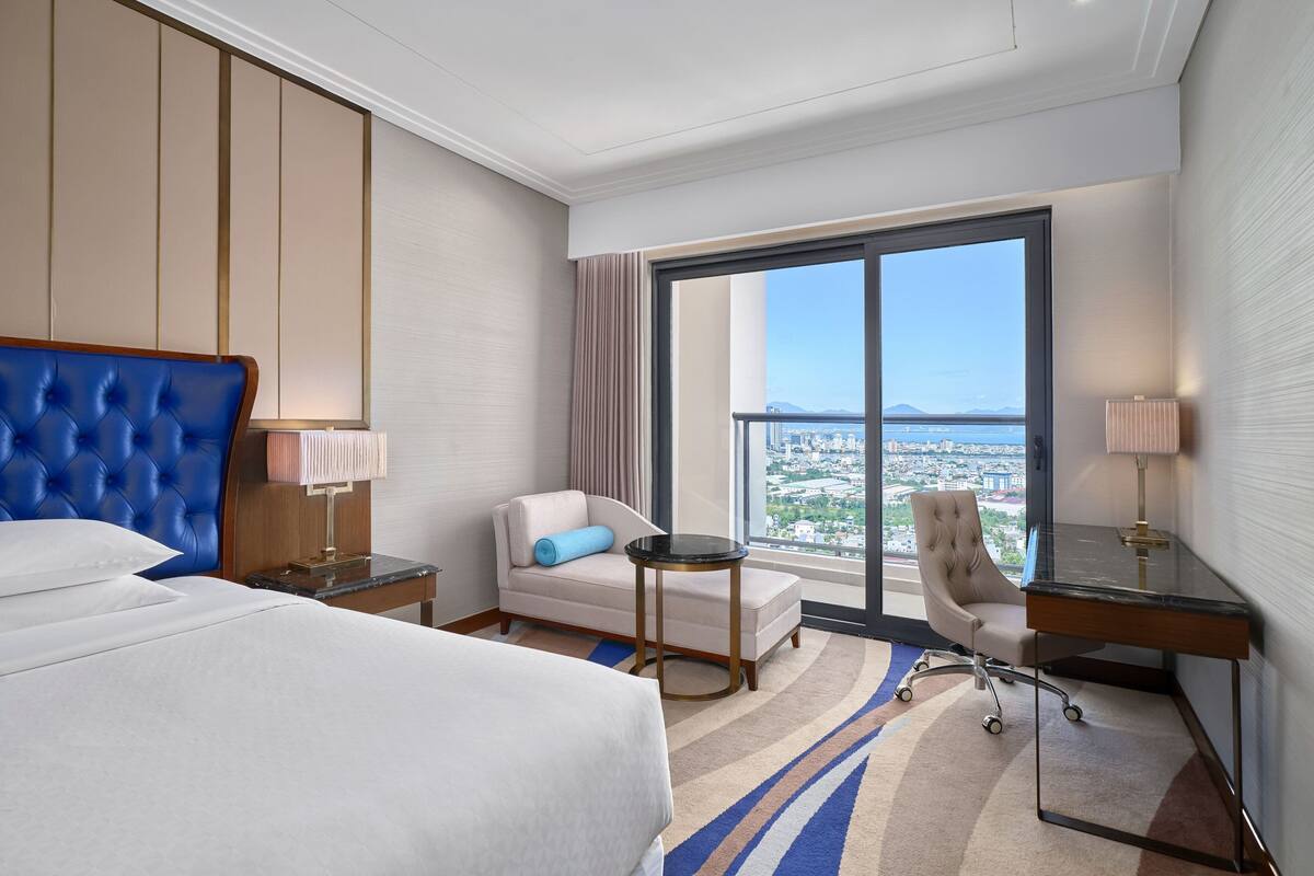 A hotel room with a large bed, a desk, a chair, and a view of the city through a large window.
