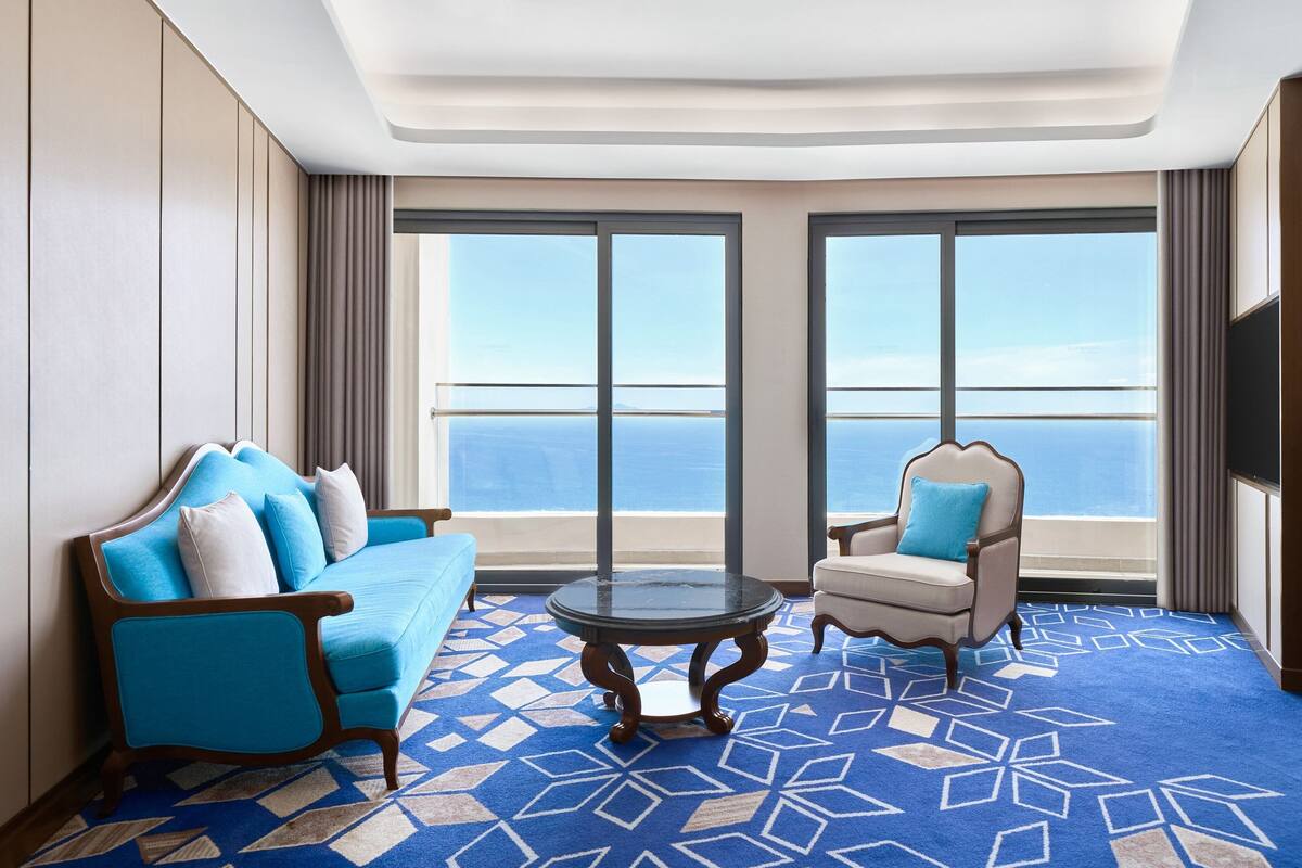 A room with a blue patterned carpet, a blue sofa with white cushions, a round table, and a white armchair with a blue cushion.