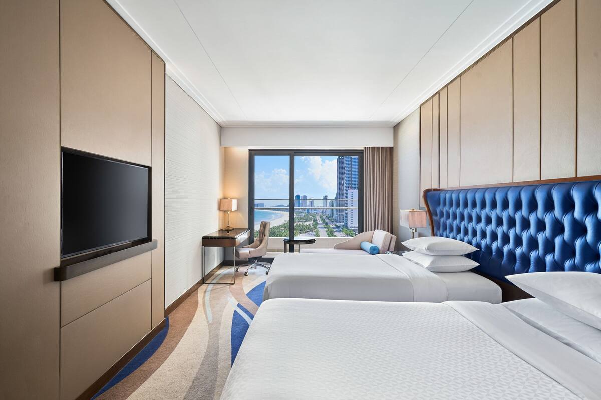 A hotel room with two beds, a TV, a desk, and a view of the city.