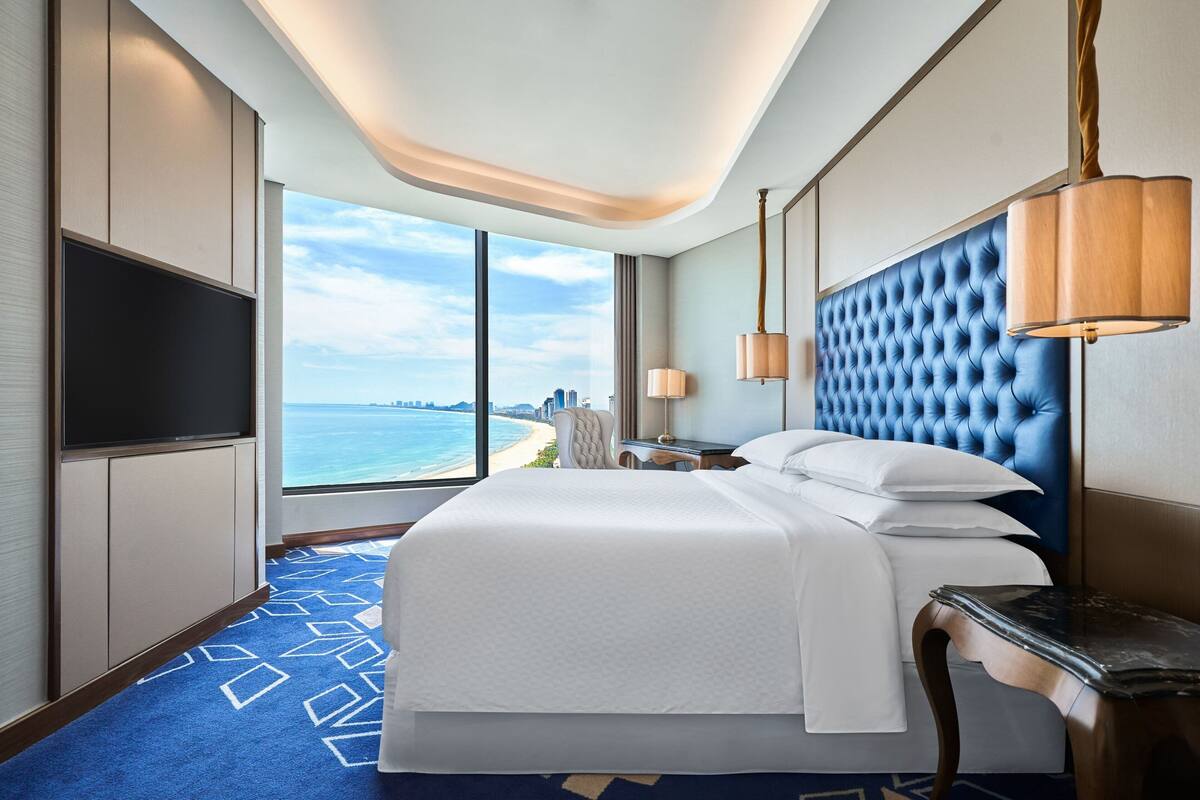 A hotel room with a large bed, a flat-screen TV, a desk, and a view of the ocean.