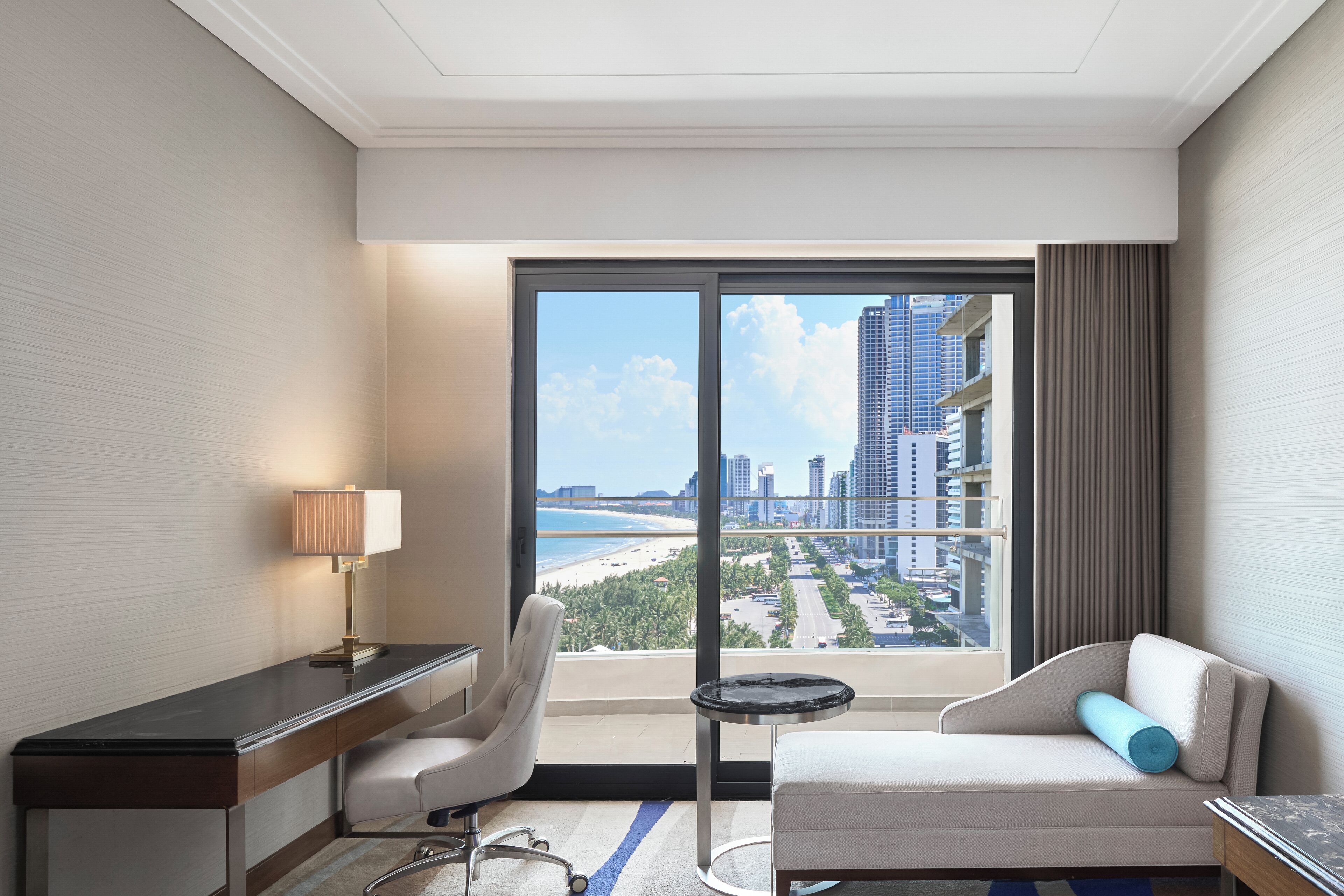 Habitación de hotel con escritorio, silla, sofá y vistas a la playa y al paisaje urbano a través de amplios ventanales.