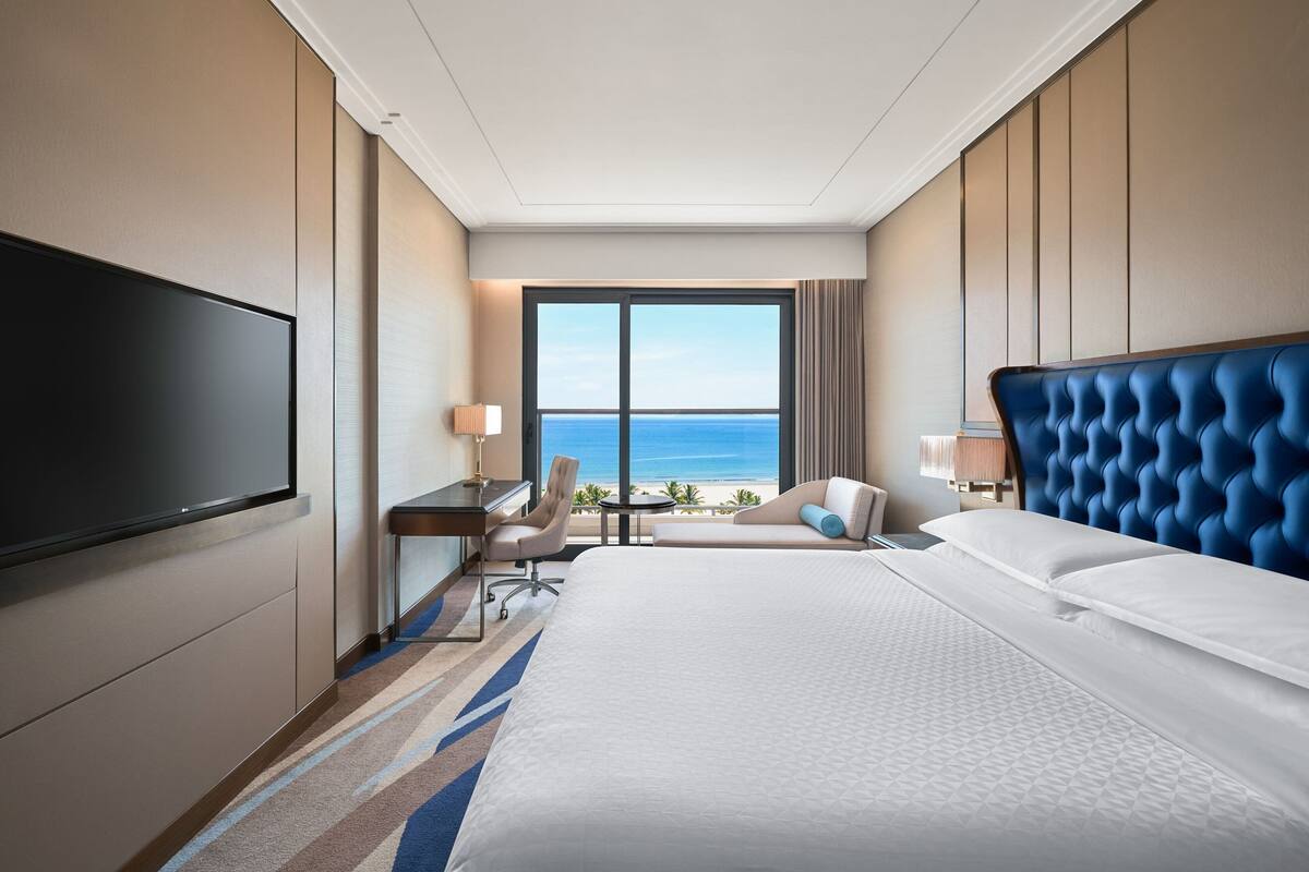 A hotel room with a large bed, a desk, a chair, a TV, and a view of the ocean.