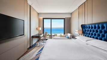 A hotel room with a large bed, a desk, a chair, a TV, and a view of the ocean.