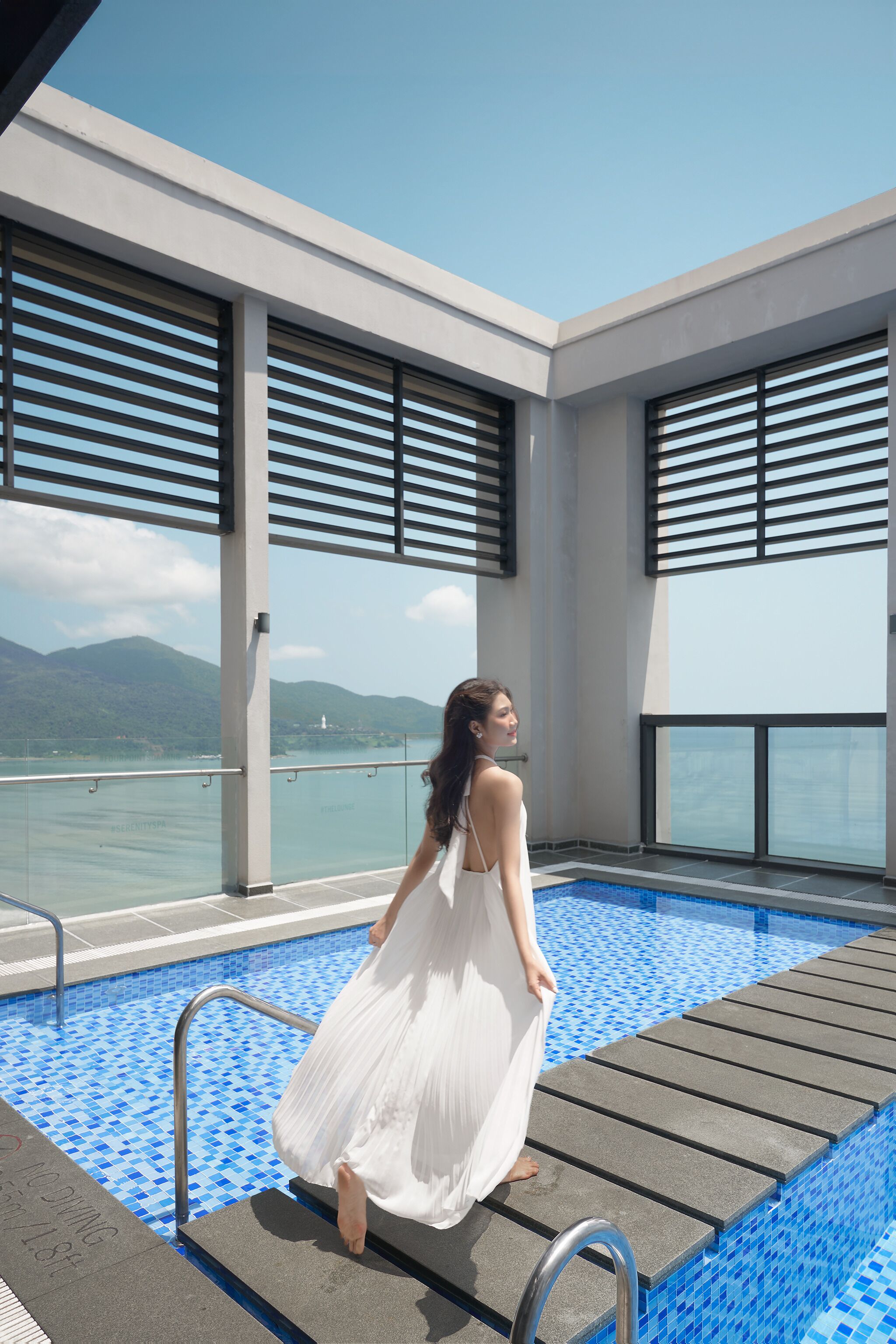 A woman in a white dress standing by a pool with a view of mountains and a body of water.