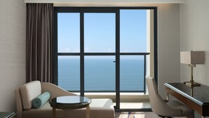 Habitación de hotel con un ventanal que ofrece vistas al océano, un sofá con un cojín, una mesita redonda y un escritorio con lámpara.