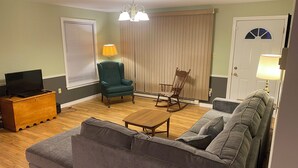 A living room with a grey sofa, a wooden coffee table, a green armchair, and a rocking chair. There is a television on a wooden cabinet and a door with a window. - Oysterbay Cottage - Fun, Beach and Food!!! License number 2100974 (Darnley)