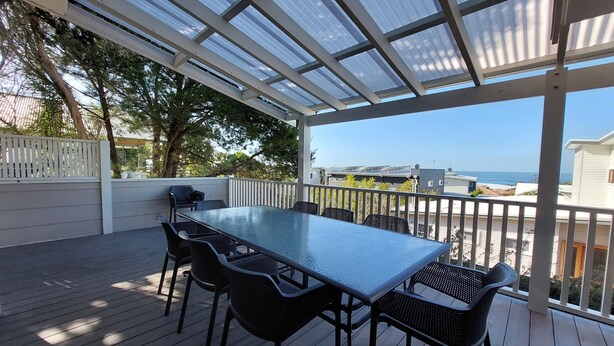 Une terrasse extérieure couverte, dotée d’une table et de chaises, qui offre une vue sur un quartier résidentiel et la mer.