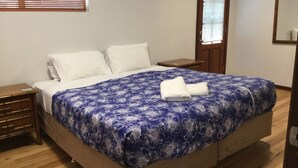Un lit bien fait, recouvert d’une couvre-lit à motifs floraux bleus et agrémenté de deux oreillers blancs, une table de chevet en bois avec un tiroir, et une porte en bois avec une fenêtre.