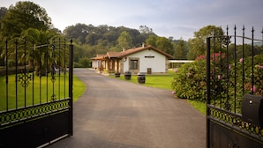 A gated entrance to a building with a paved driveway leading to a white house with a tiled roof. - El Puentuco (Villacarriedo)