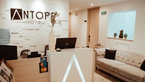 A hotel reception area with a modern design, including a reception desk, a large illuminated 'A' sign, a sofa, and a window with plants. - Antope - Apartaestudios (Cali)