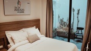 A hotel room with a large bed, a wooden headboard, a view of a balcony with plants, and a wall-mounted artwork. - Antope - Apartaestudios (Cali)
