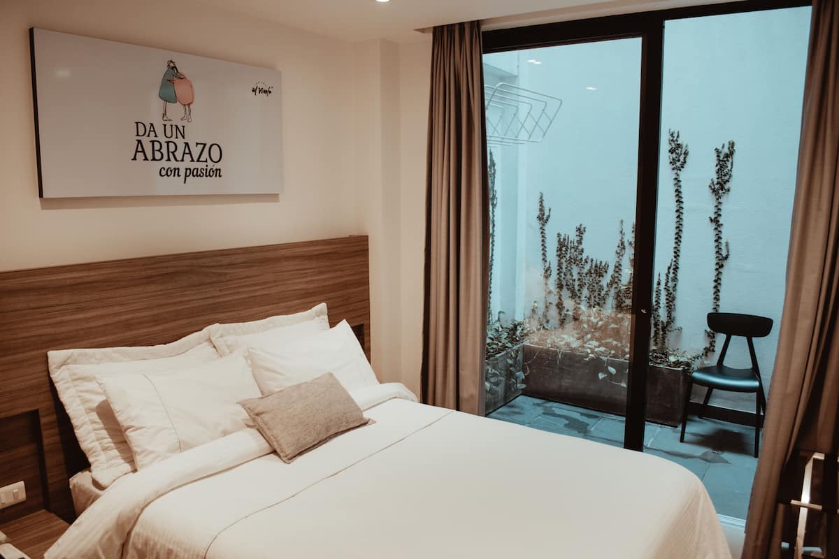 A hotel room with a large bed, a wooden headboard, a view of a balcony with plants, and a wall-mounted artwork.