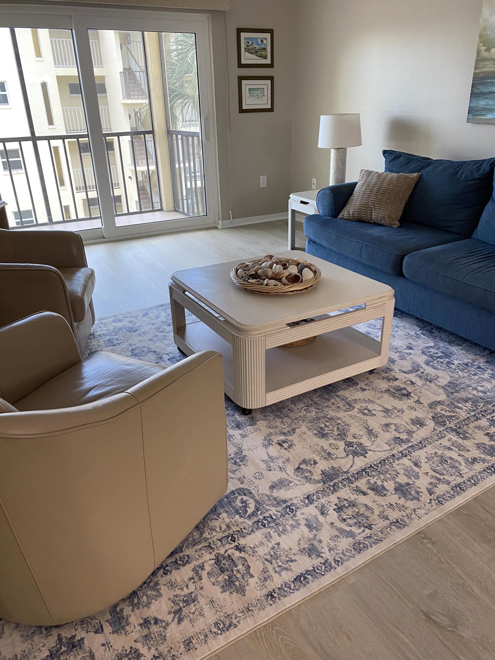 A living room with a sofa, a chair, a coffee table with a basket, a patterned rug, and a view of a building through a glass door.