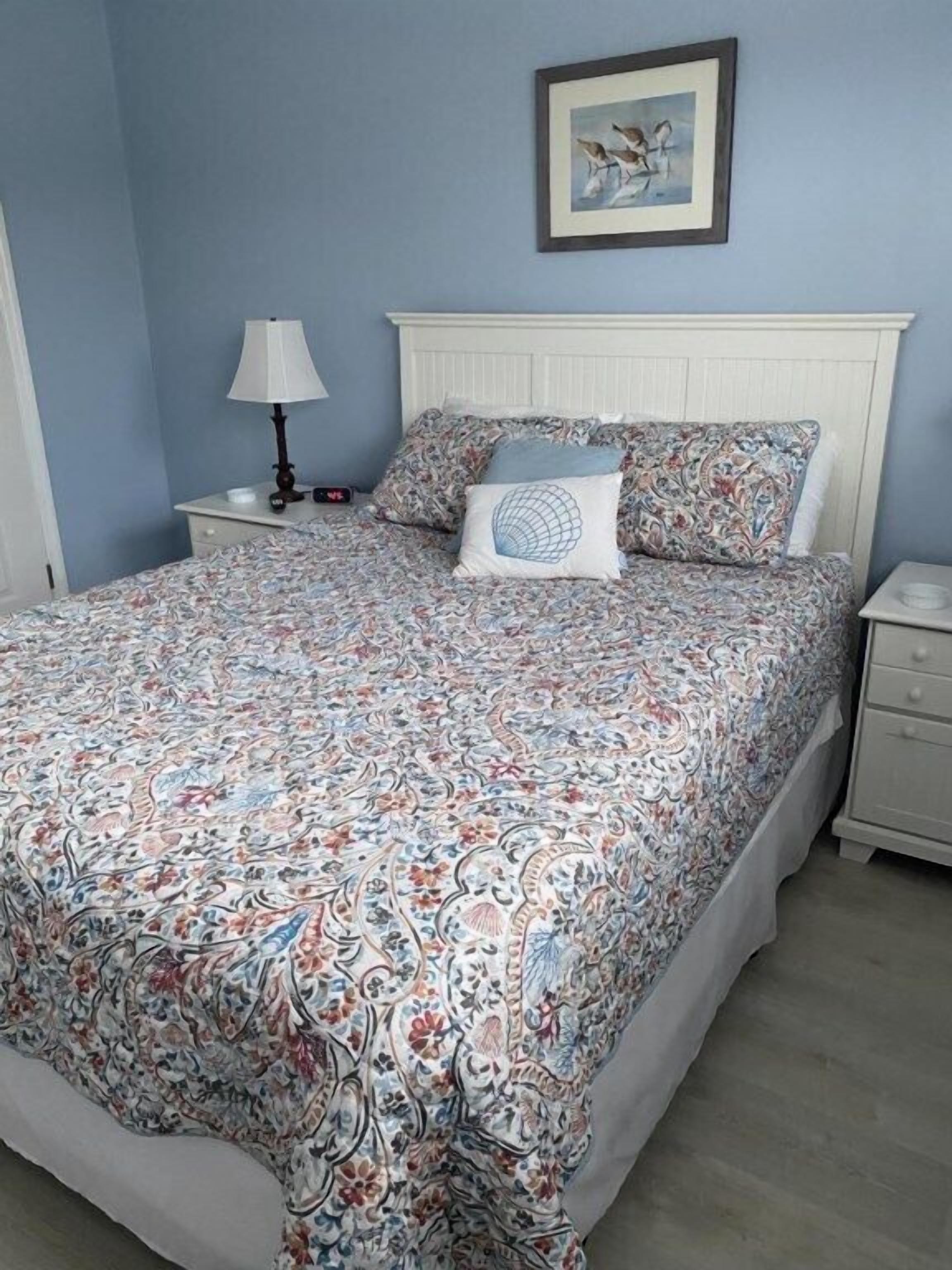 Une chambre avec une couvre-lit à motifs, une table de chevet avec une lampe, un tableau encadré au mur et une commode blanche.