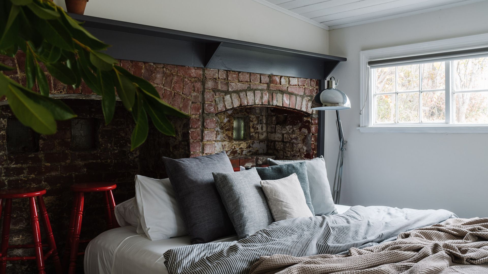 Une chambre avec une cheminée en briques, un lit avec des oreillers, un tabouret rouge et une lampe sur pied.