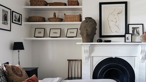 A room with a fireplace, shelves with baskets and framed pictures, a lamp, and a bed with pillows. - Piccadilly House Daylesford ~ Walk to main street! (Daylesford)
