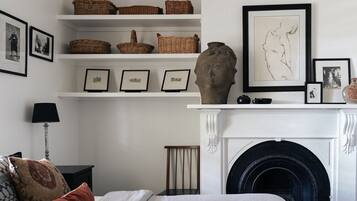 A room with a fireplace, shelves with baskets and framed pictures, a lamp, and a bed with pillows.