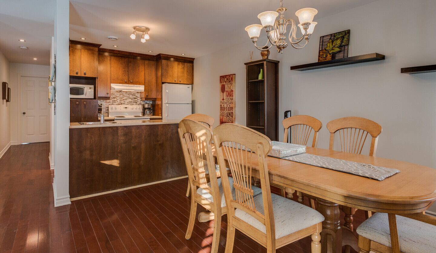 A dining area with a wooden table and chairs, a kitchen in the background, and a chandelier.
