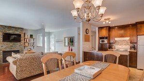 A modern living room with a fireplace, a dining area with a chandelier, and a kitchen with wooden cabinets and a microwave. - Condo Magog entre Ville et Nature (Magog)
