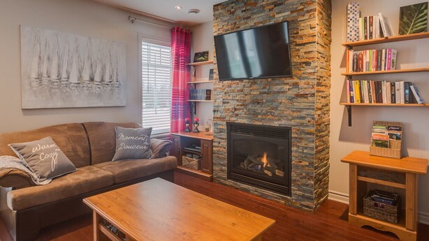 A living room with a stone fireplace, a flat-screen TV mounted above it, a brown sofa with decorative pillows, a wooden coffee table, and bookshelves filled with books. - Condo Magog entre Ville et Nature (Magog)