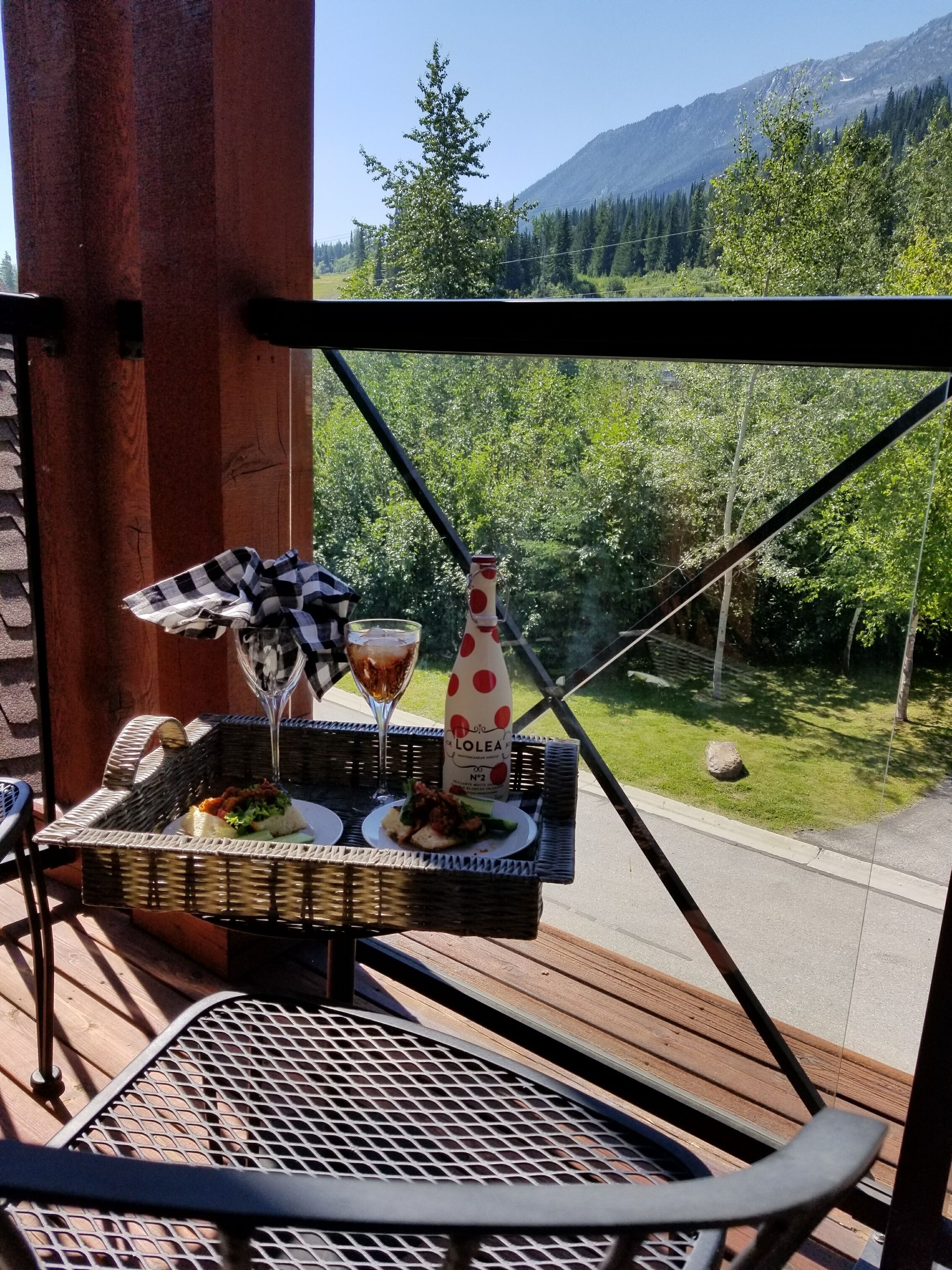 Un balcón con una mesa de mimbre dispuesta con comida y bebidas, con vistas a una zona boscosa y montañas.