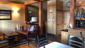 A cozy living room with a brown leather sofa, a coffee table, and a dining table with chairs. There is a kitchen area with a stove, a sink, and a clock on the wall. - Slope Side  Luxury Open Beam Townhome Private Hot Tub - True - Ski-in-out (Golden)
