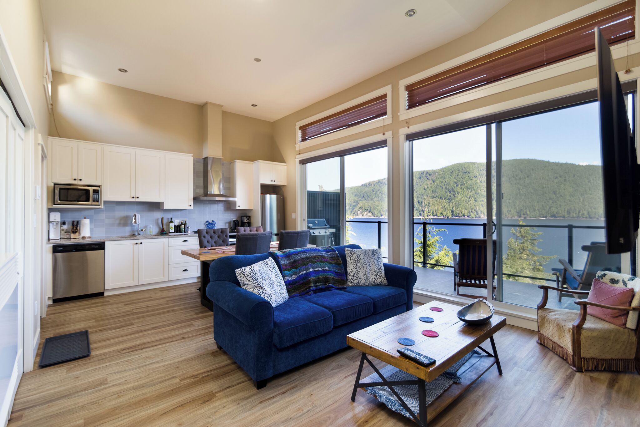 A modern living space with a kitchen, a blue sofa, and a large window offering a view of a forested hill.