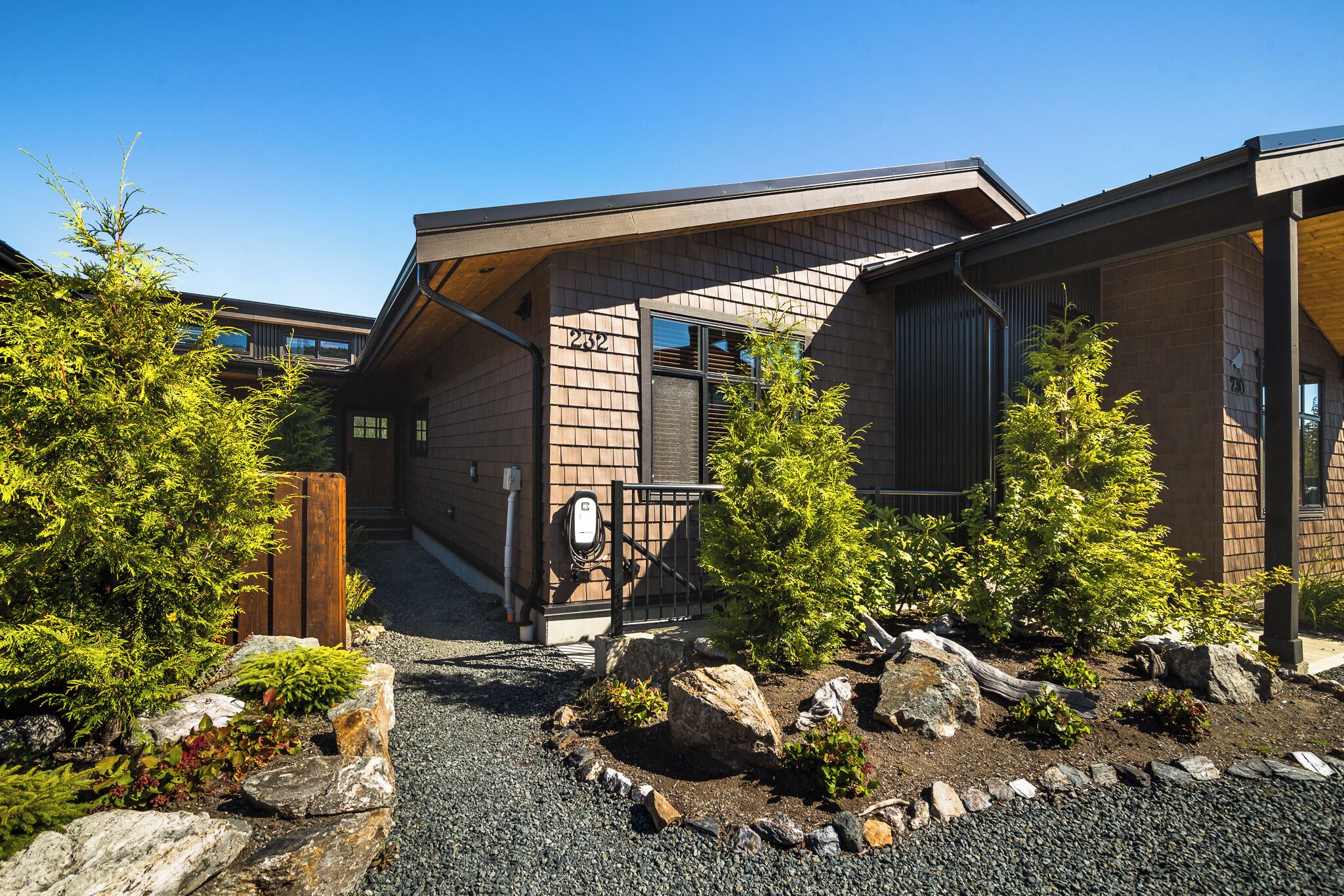 A modern building with a sloped roof, a wooden fence, and a gravel pathway with rocks and small trees.