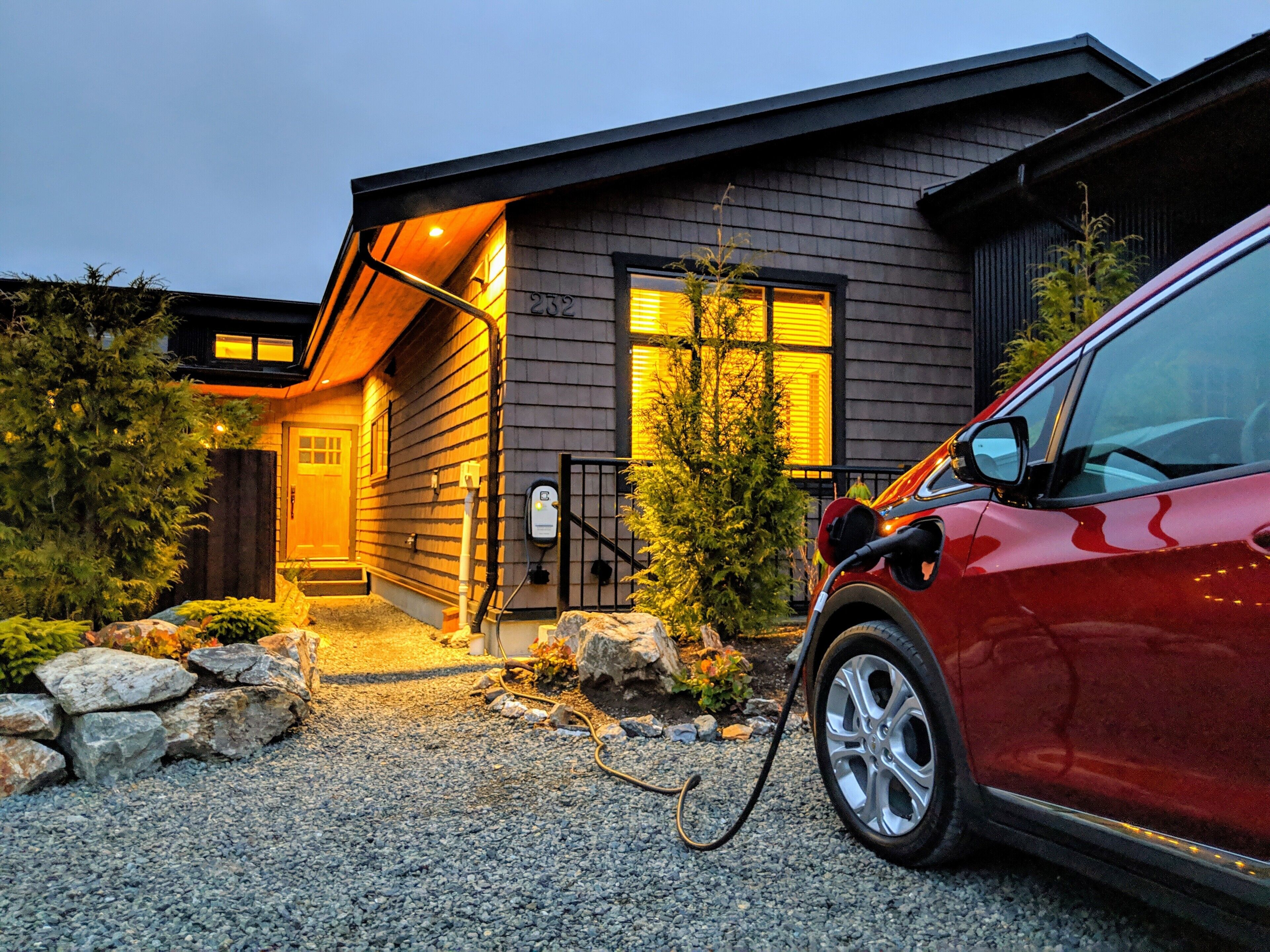 Uma casa moderna com entrada por uma calçada, uma estação de carregamento para carro e uma pequena árvore.