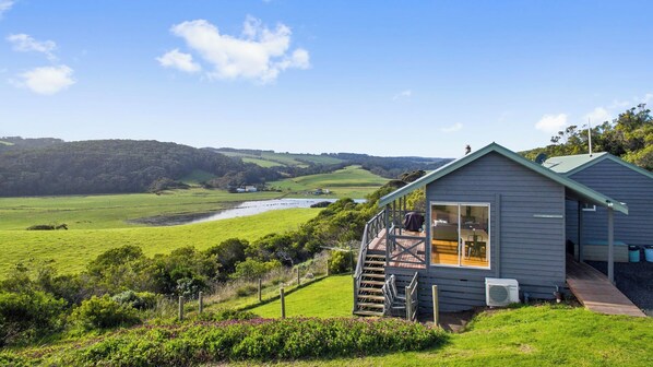 A small cabin with a deck, overlooking a river and green fields. - Rivernook Beach Cottage (Johanna)